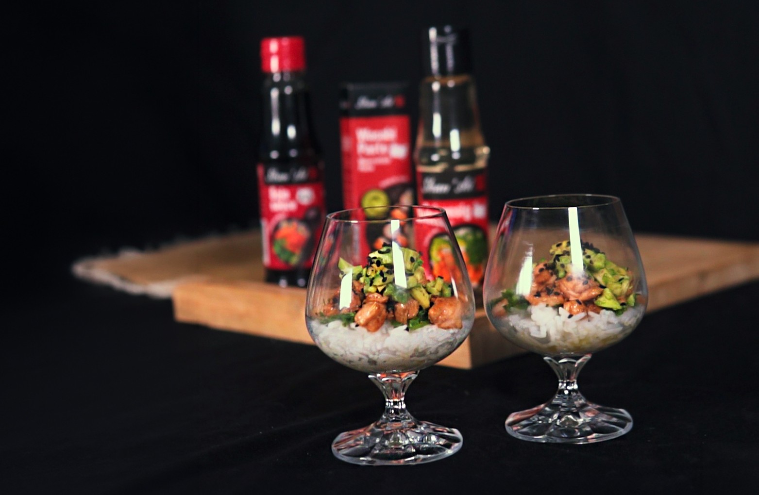 Hotový sushi šalát s ryžou, lososom, avokádom a rukolou v dvoch sklenených pohároch; v pozadí prísady Shan’shi.